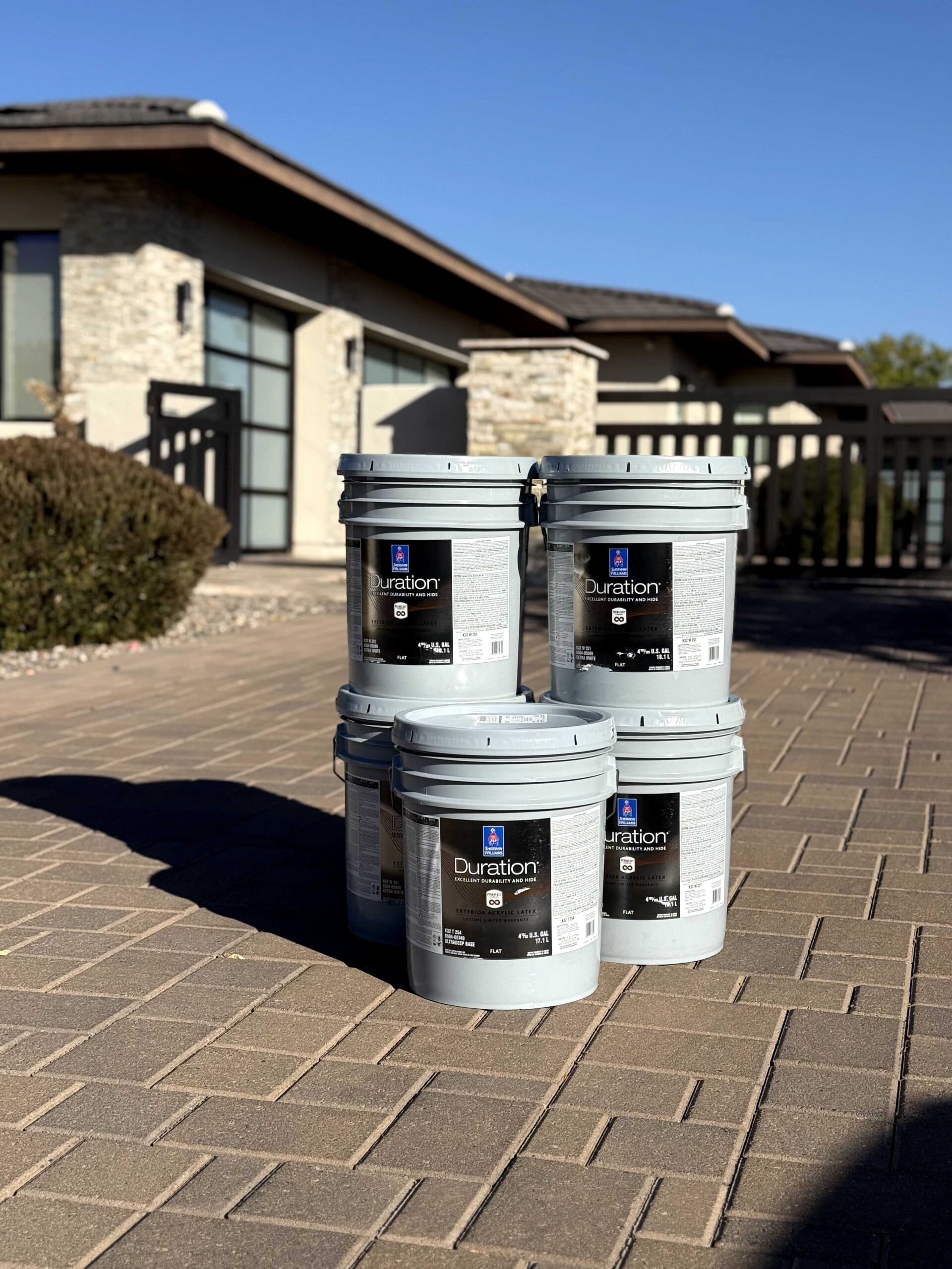 Five large gray buckets of Sherwin-Williams Duration exterior paint stacked on a paved driveway in front of a residential house.
