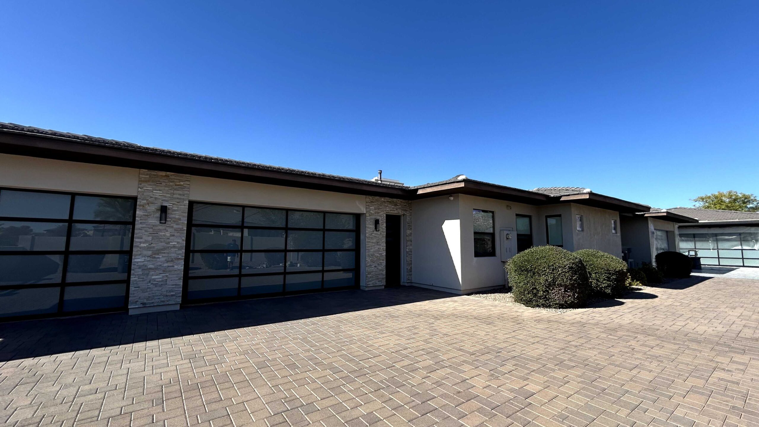A modern, light-colored stucco house with two large, contemporary garage doors featuring frosted glass panels and dark frames. The house has stone veneer accents on the pillars between the garage doors and a light brown tiled roof. The driveway is made of patterned pavers, and the sky is clear and blue.