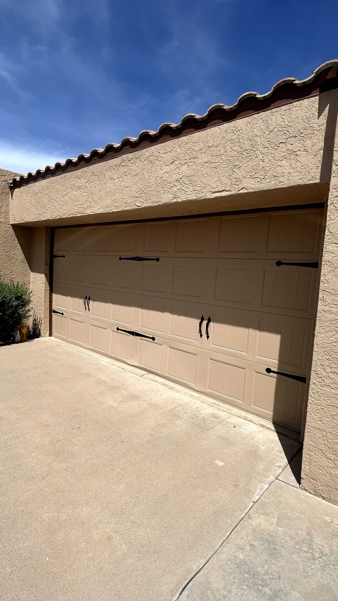A close-up view of a light brown garage door with decorative black hardware, including hinges and handles, on a stucco house. The house has a tiled roofline. The sky is blue with a few wispy clouds.