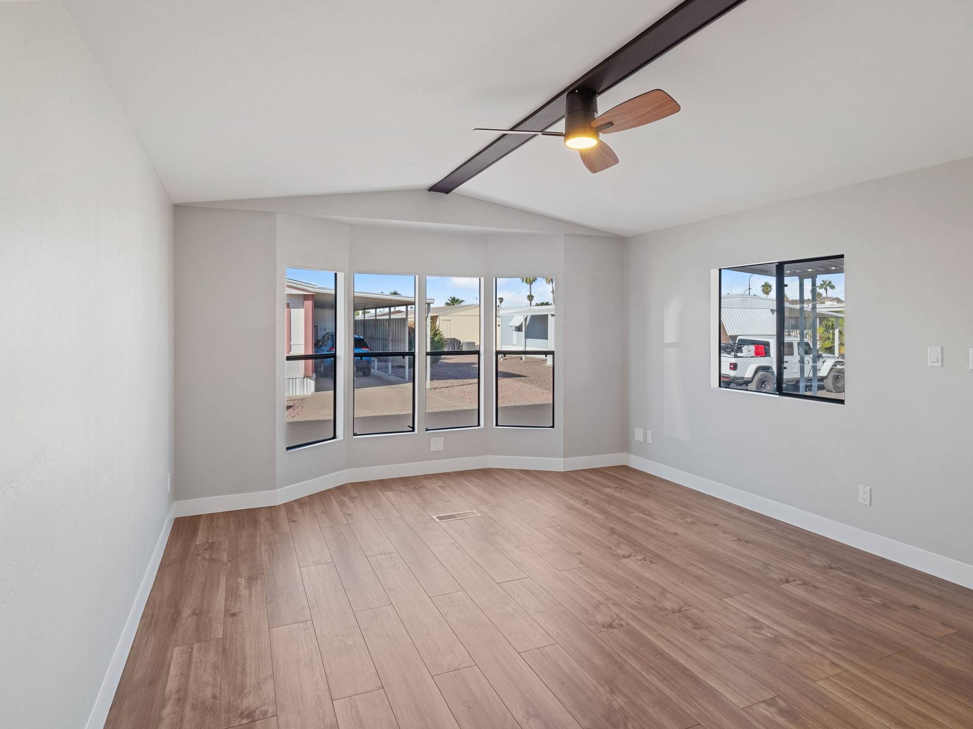 An empty, freshly painted living space with light walls, wood-look flooring, a bay window, and a ceiling fan with a dark beam.